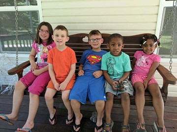 6 kids sitting on a bench on a porch during the day.